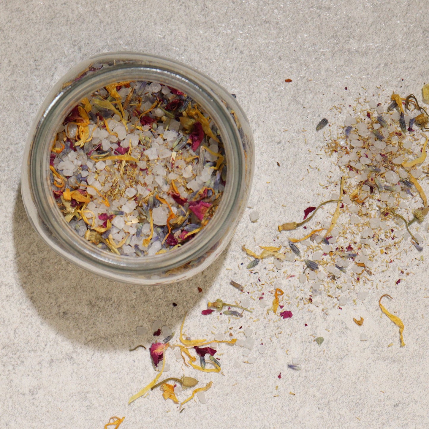 Blumiges Badesalz mit Blüten – geöffnetes Glasgefäß, Draufsicht mit verstreuten Blüten auf hellem Grund
