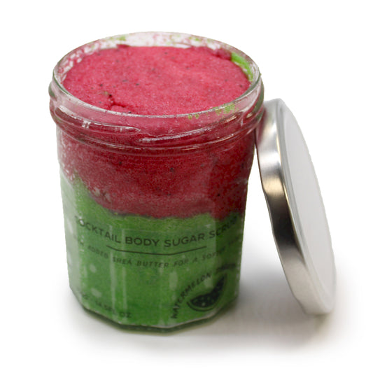 Zuckerkörperpeeling mit Wassermelonen-Daiquiri-Duft, Peeling-Kristalle in rosa und grüner Farbgebung, in durchsichtigem Behälter mit silberfarbenen Deckel, der abgeschraubt an das Glas gelehnt steht.