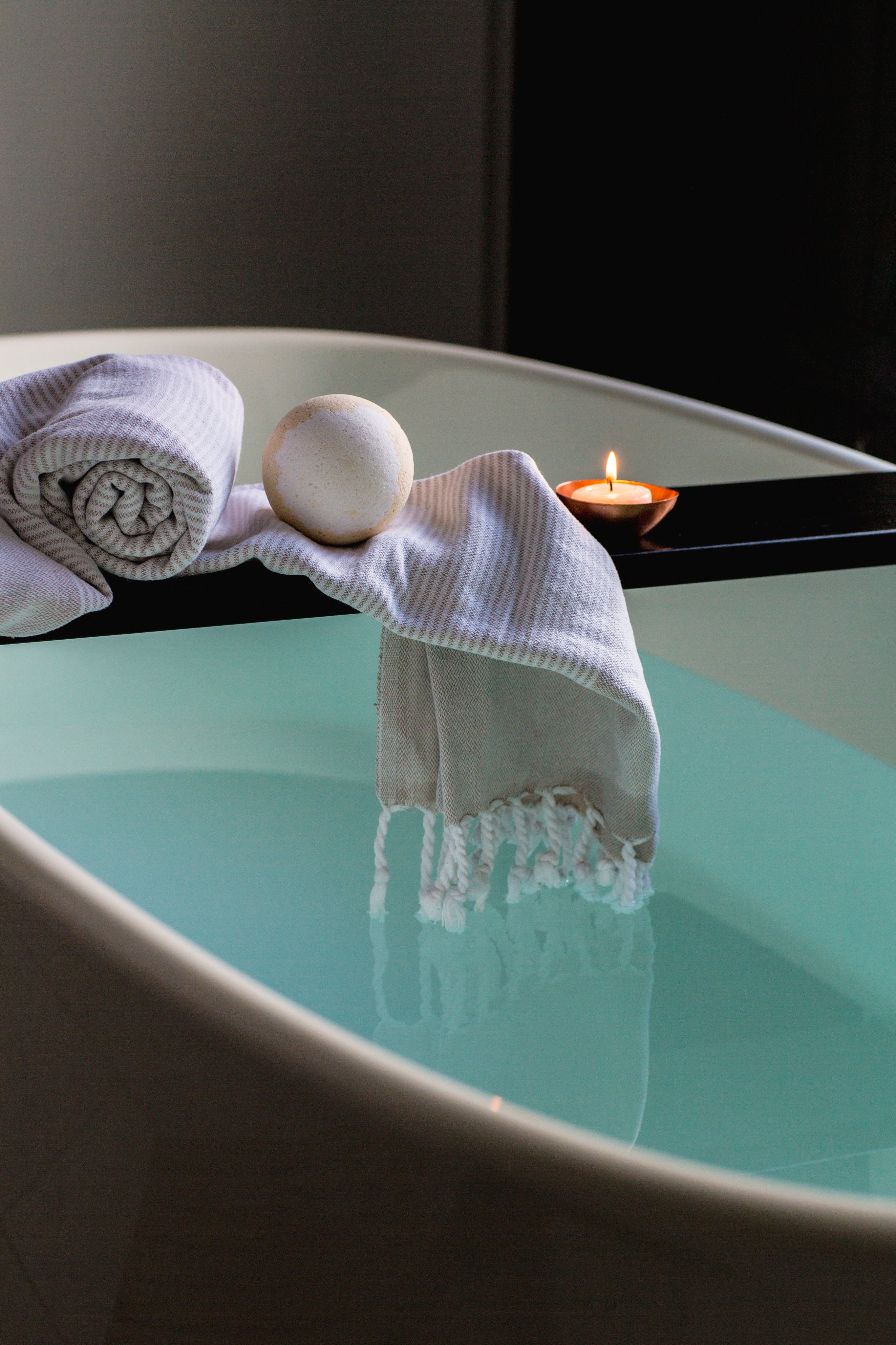Badewanne mit türkisfarbenem Wasser, Badebombe, gerolltem Handtuch und brennender Kerze auf Wannenablage – Baderitual als Auszeit